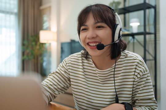 Smiling young Asian student wearing a headset joins an online class from home, using a laptop for video communication, remote learning, and digital education.