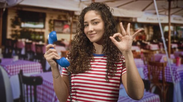 Woman holding blue vintage telephone handset, making ok sign with hand in a street cafe with red checkered tables; playful confidence.