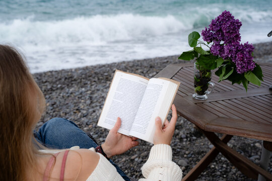 Back view of a young woman reading a printed book while sitting by the ocean. Quiet seaside moment with focus on mindfulness and digital detox.
