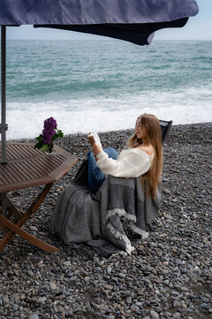 Woman relaxing with a book on a rocky seaside beach, wrapped in a cozy blanket while enjoying a quiet moment by the ocean. Calm lifestyle, mindfulness and outdoor leisure.