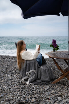 Young woman enjoying a printed book on a pebble beach with ocean waves in the background. Peaceful travel and mindful recreation.