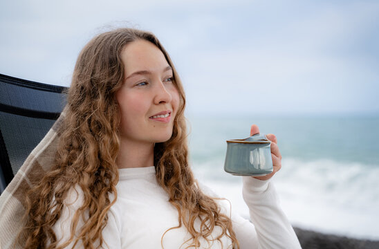 Curly woman enjoying a warm drink beside the ocean. Calm seaside atmosphere, slow living and mindful wellbeing concept.