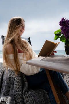Happy woman enjoying a paper book while sitting outdoors on a pebble beach. Slow living, digital detox and peaceful seaside atmosphere.