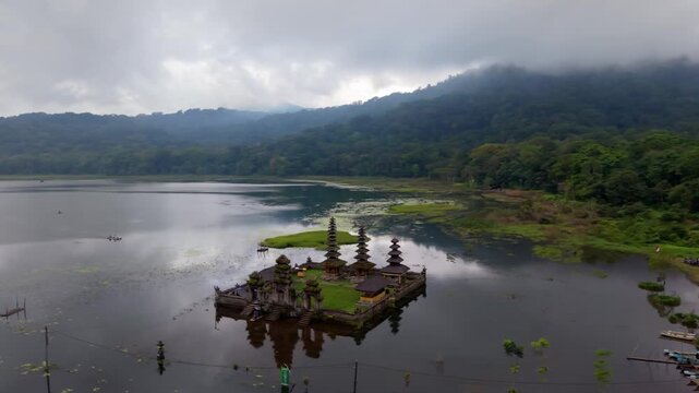 Aerial view of Ulun Danu Tamblingan Temple on Lake Tamblingan surrounded by mountains and forest under a cloudy sky in Bali, Indonesia