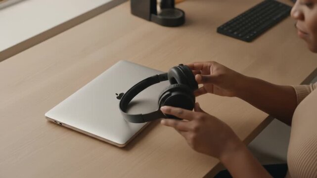 An African American woman sits at a desk, carefully inspecting a pair of black over-ear headphones next to her closed laptop in a minimalist workspace.