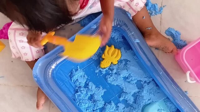 Close-up video of a child's hand playing with blue kinetic sand in a plastic tray. The toddler uses a yellow shovel and pink crab mold for sensory play.