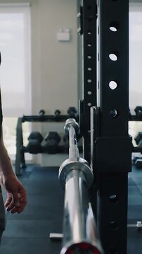 Vertical video: Entering man in gym wear standing by rack in small gym, preparing lift with barbell