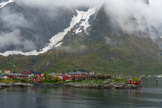 Red cabins sit on the shore of a fjord under cloudy skies in Reine, Lofoten archipelago, Norway