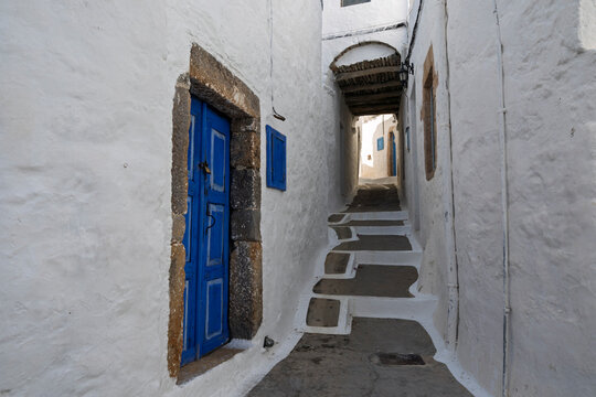 Narrow pathway leads to blue door in Chora, featuring white stone walls and steps in the Dodecanese Islands, Greece