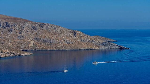 Sailing boats navigate the calm waters near rocky shores of Leros Island in Greece during a bright day