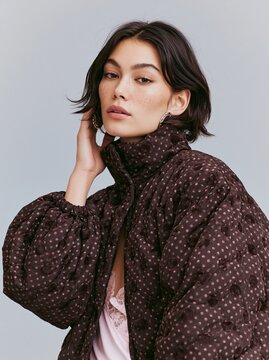Woman wearing a dark brown polka dot high neck jacket poses with one hand on her neck, showcasing a stylish look against a soft, neutral background