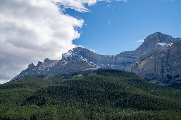 Fototapeta premium Layered limestone cliffs rise above dense evergreen forest under dramatic skies at Lake Minnewanka in Banff National Park, Alberta, Canada.
