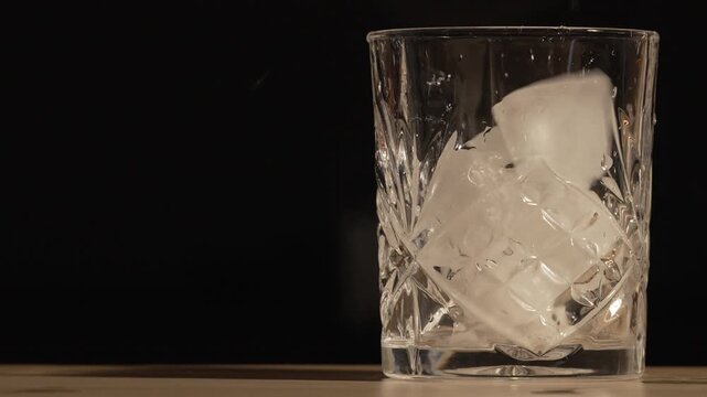 Ice Cubes Dropping into an Empty Crystal Whiskey Glass on a Wooden Bar Table with Dark Background