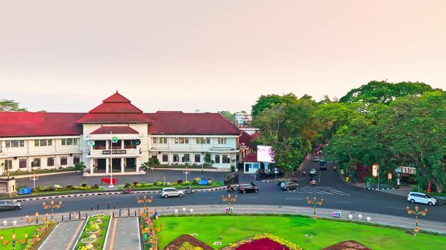 Golden hour bathes historic building and central monument. Circular pond reflects sky and structure. Lush greenery surrounds the scene. Distant trees frame the peaceful vista
