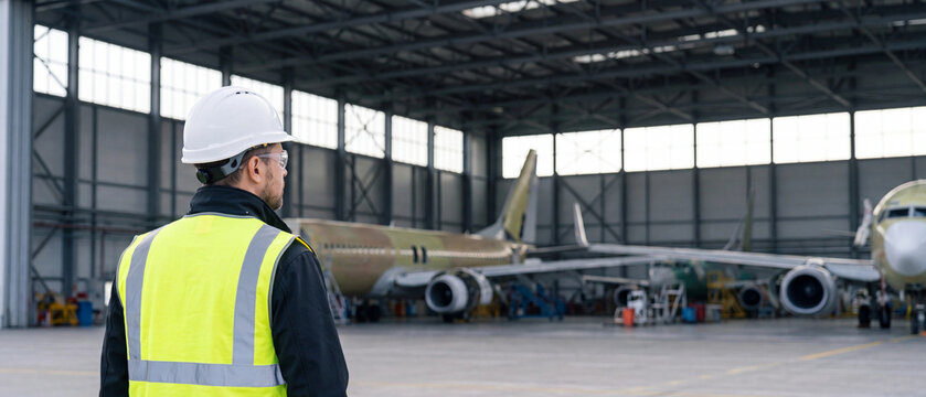 A male aircraft maintenance engineer in a hard hat and highvisibility vest standing in a large airplane hangar
