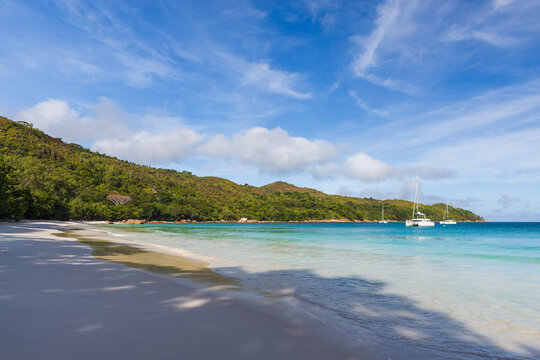 A tranquil tropical beach with white sand, clear turquoise water and an anchored yacht framed by green hills and a bright blue sky with soft white clouds. Seychelles, Anse Lazio