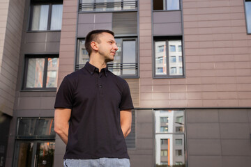 Naklejka premium Caucasian man standing outdoors in front of a modern residential building. Young guy in a black polo shirt looking away. Urban lifestyle, professional portrait and city living concept.