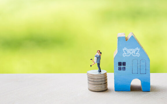 Miniature couple on coin stack with miniature house over blurred natural green lawn background, love and romance concept
