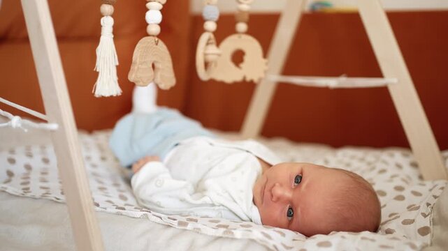 Sweet infant boy wearing cozy pajamas lies on a play mat under a montessori style wooden activity gym, looking around and developing his senses in a comfortable nursery setting