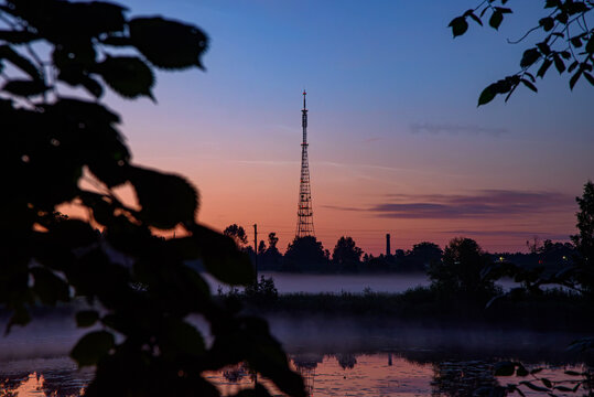 Misty sunrise over a calm lake with a tall radio tower, soft reflections, silhouetted trees, and colorful dawn sky.