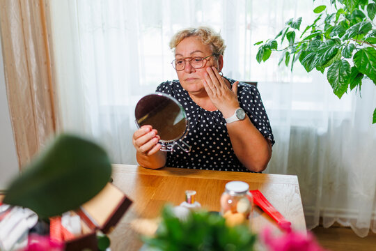 Senior woman examining facial aging and cheek skin in handheld mirror.