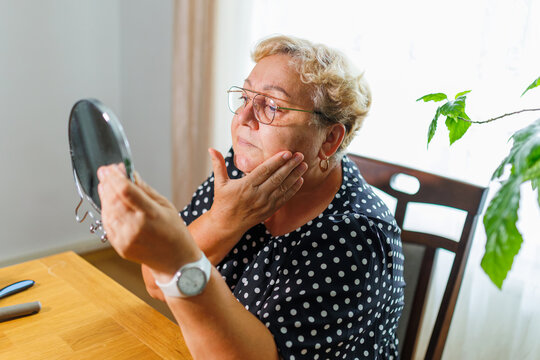 Senior woman checking facial skin texture with handheld mirror indoors.