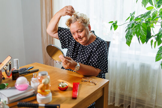 Mature female touching head and examining grey hair roots at wooden table.