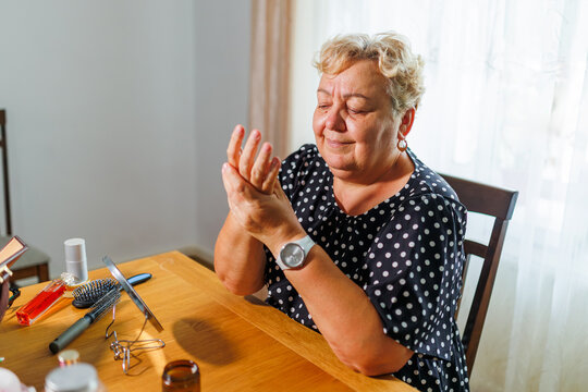 Senior woman rubbing moisturizing hand cream at a wooden living room table.
