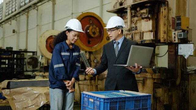 Asian manager and female worker in hard hats discuss and criticizing quality control of a machined metal part using a laptop. They stand near heavy machinery in a large industrial manufacturing plant 