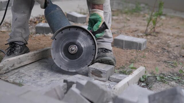 Close-up outdoor construction scene with worker using angle grinder on concrete block near center-right, green glove guiding tool beside dusty ground, curb stones, weeds, muted