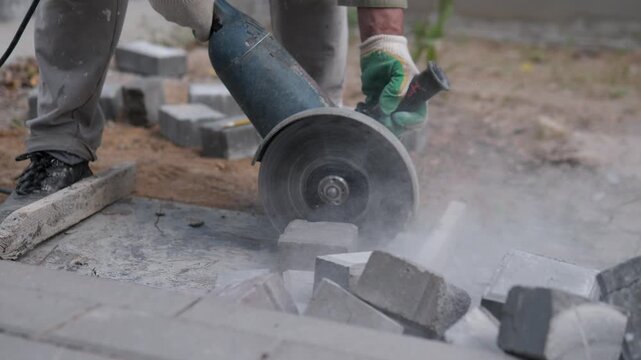 Worker in gloves cuts concrete pavers outdoors with handheld circular saw, dust rising around scattered blocks, rough stone surface, boots, and unfinished walkway under soft