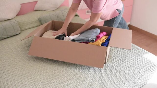 An organized Caucasian woman neatly placing folded clothing into a cardboard box on a bed, getting ready for moving house or decluttering her home wardrobe