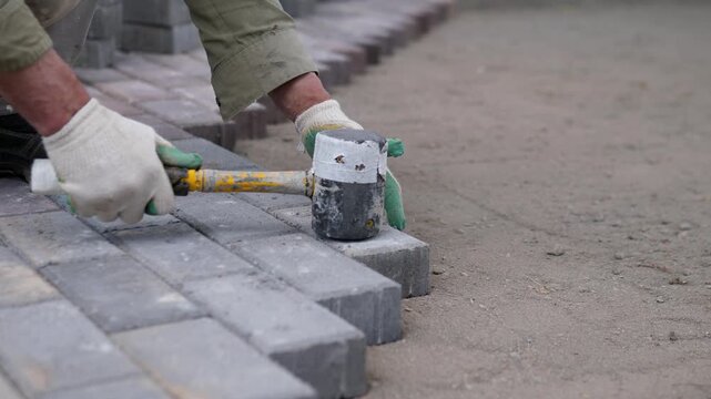 Close view shows construction worker setting gray paving stones along outdoor curb line with gloved hands and mallet, forming clean alignment across sandy ground under muted