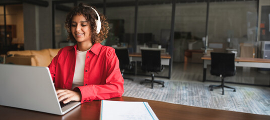 Naklejka na ściany i meble Young indian woman with curly hair working on laptop computer at desk in modern office or home workspace, wearing shirt and smiling. Latin or muslim arab student girl studying. Web banner, copy space