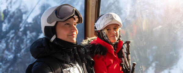 Two female skiers laugh together inside mountain gondola, holding poles beside snowy alpine scenery. Joyful winter moment captures friendship, ski fun, happiness, travel, and holiday energy © Kyryl Gorlov