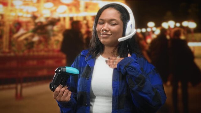 Young woman holding a game controller with hand to chest, wearing headset and septum ring in a street fair at night framed by carousel lights and a crowd; contentment.