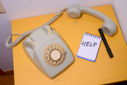 Close-up of an old rotary phone on a yellow wooden table and a small notepad with the word help written on it.