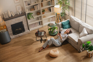 Obraz premium Overhead view of woman relaxing on floor with smartphone in cozy living room, fireplace and bookshelf background, laptop on table, warm home interior lifestyle scene with copy space