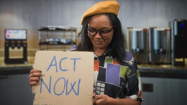 Young african american woman holding banner act now points finger to chest in building; urgent activism solidarity.