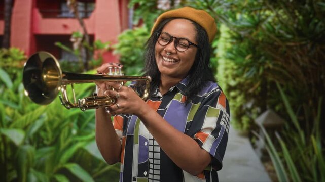 Young woman holding trumpet with fingers on valves, smiling with closed eyes in building courtyard; joy creativity.