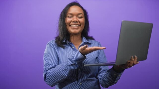 Young african american woman holds laptop in left hand and gestures palm up with right in studio; optimism presentation.