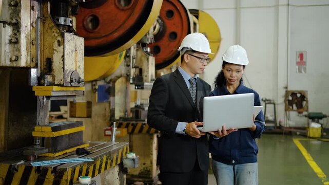 Asian male engineer and black female factory worker wearing hard hats discuss manufacturing process using a laptop near a large industrial press machine, inspecting a metal part for quality control.