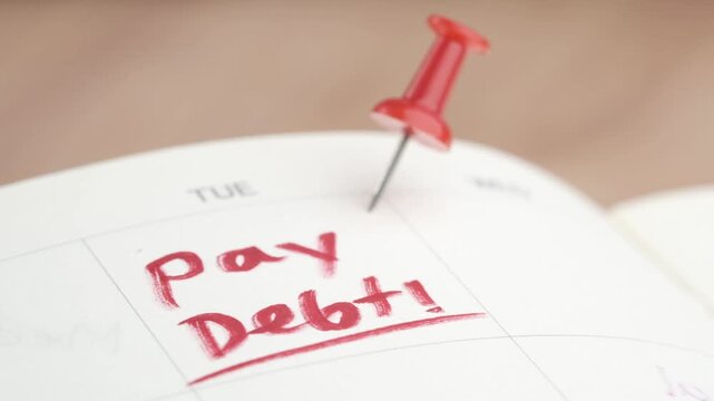 Close-up cinematic footage of calendar with red thumbtack pinned on tuesday with handwritten red text reading 'pay debt!'