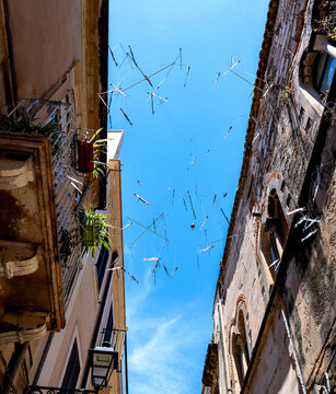  Small decorative silver-coloured metal fish suspended between historic buildings glisten against Sicily&rsquo;s deep blue sky