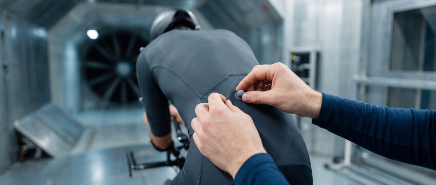 Cyclist undergoing aerodynamic wind tunnel testing with technician attaching sensor to skinsuit