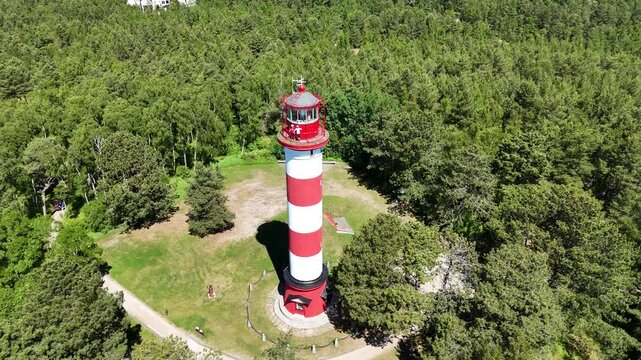 High angle aerial panoramic view of Nida lighthouse on the Curonian Spit, Lithuania. Drone shot of the red and white striped beacon surrounded by lush green pine forest in summer.