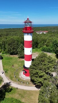 Aerial vertical video of Nida lighthouse on the Curonian Spit, Lithuania. Drone orbit shot of the red and white striped beacon in green pine forest with Baltic sea on background.