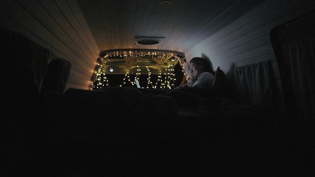 Woman and her daughter sit together in a camper van and watch a movie on a laptop. The lights create a cozy setting.