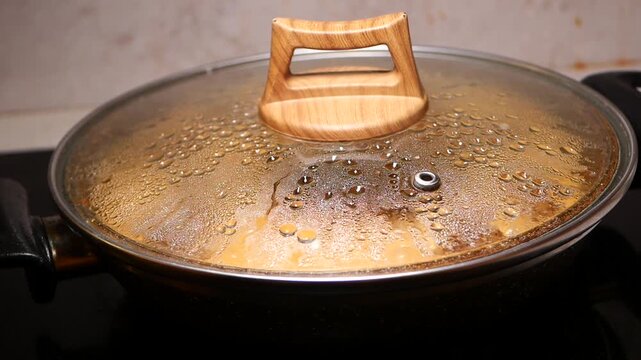 Cooking food in frying pan with glass lid covered with condensation and water droplets on induction stove