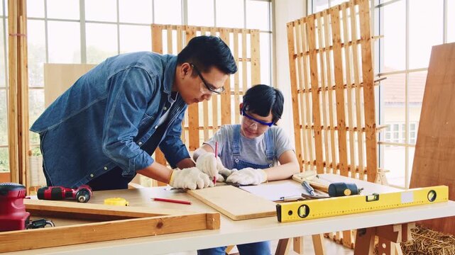 Happy Asian father and son carpentry wearing safety gloves. Father teaches his son to measure wood by measuring tape and marking with a pencil. Home workshop and Carpentry family concept.